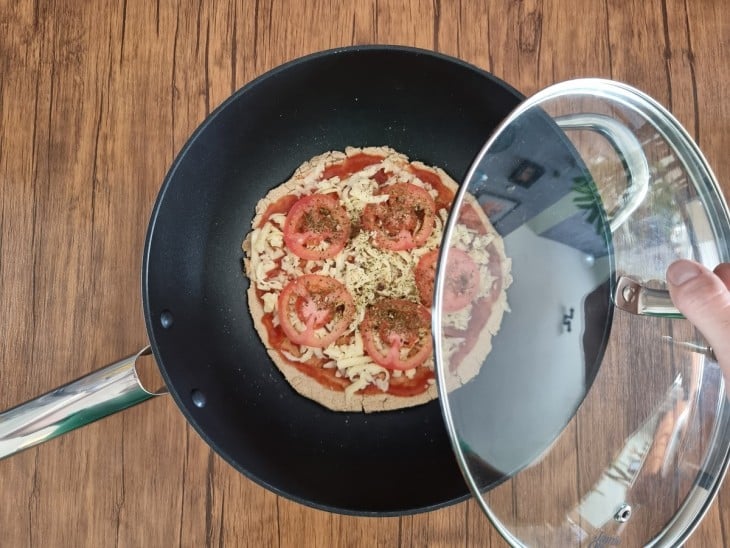 Recheios de tomate, queijo e molho sobre pizza, dentro de frigideira preta com tampa de vidro.