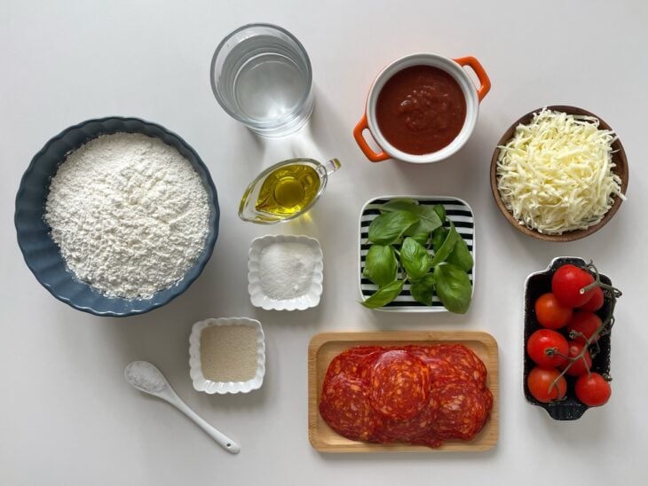 Bancada organizada com farinha, queijo mussarela ralado, rodelas de pepperoni e tomates-cereja em potes.
