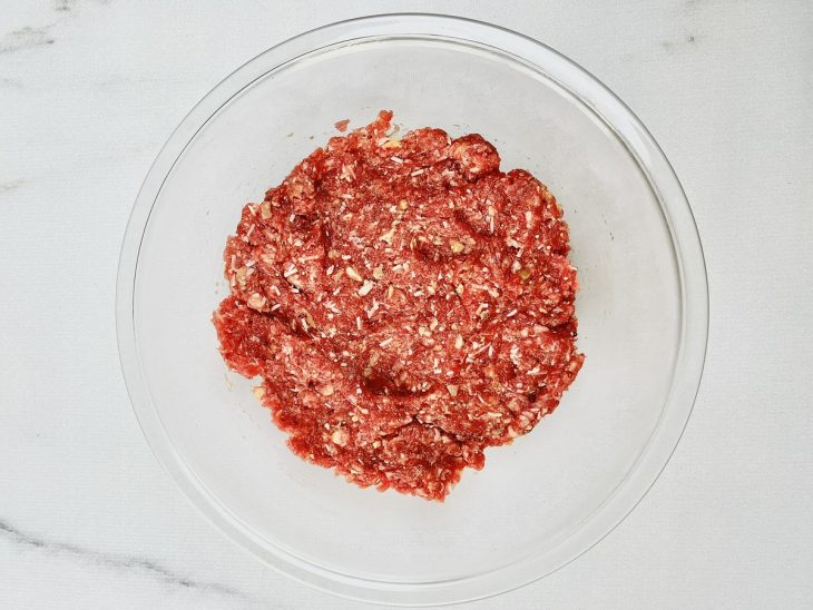Carne moída, ovo, mostarda dijon, parmesão ralado, farinha de pão, pimenta, sal e molho inglês misturados em uma tigela de vidro.