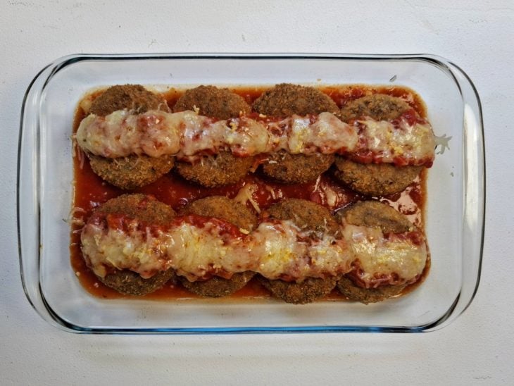 Polpetones acomodados em um recipiente de vidro com fundo forrado por molho de tomate, e parmesão salpicado por cima.