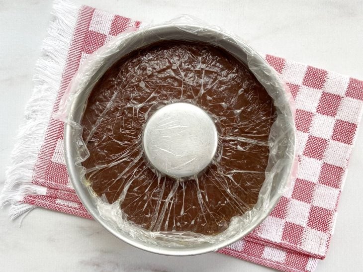 Uma forma contendo a massa do pudim de chocolate, coberto com plástico tocando na superfície o doce.