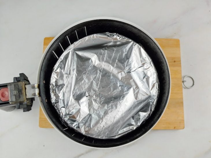Forma de pudim coberta com papel alumínio dentro do cesto da airfryer.