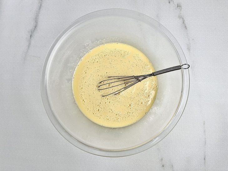 Ingredientes batidos com auxílio de um fouet em uma tigela redonda de vidro.
