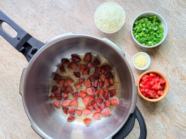 Na panela de pressão, a calabresa refogada, ao lado, o tomate, a cebola, o alho e o pimentão.