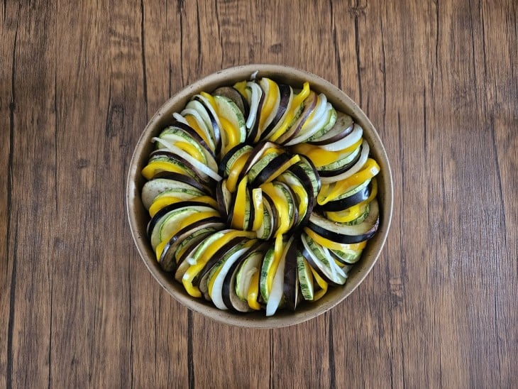Uma assadeira redonda com fatias intercaladas de berinjela, abobrinha, pimentão amarelo e cebola.