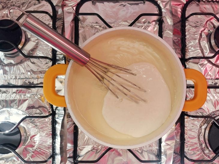 Um fouet e uma panela com leite condensado, amido de milho, leite em pó, leite, creme de leite e manteiga.