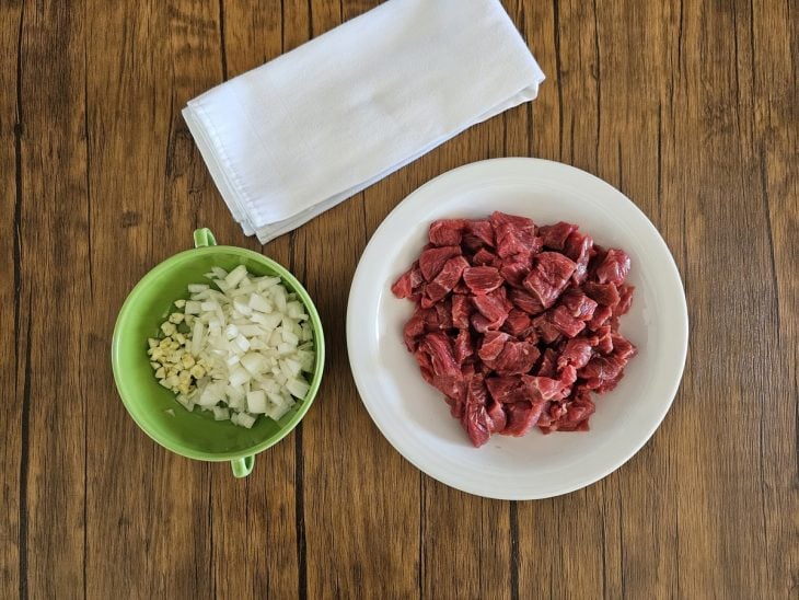 carne para o risoto cortada em pedaços pequenos em um prato e cebola e alho picados em um pote pequeno