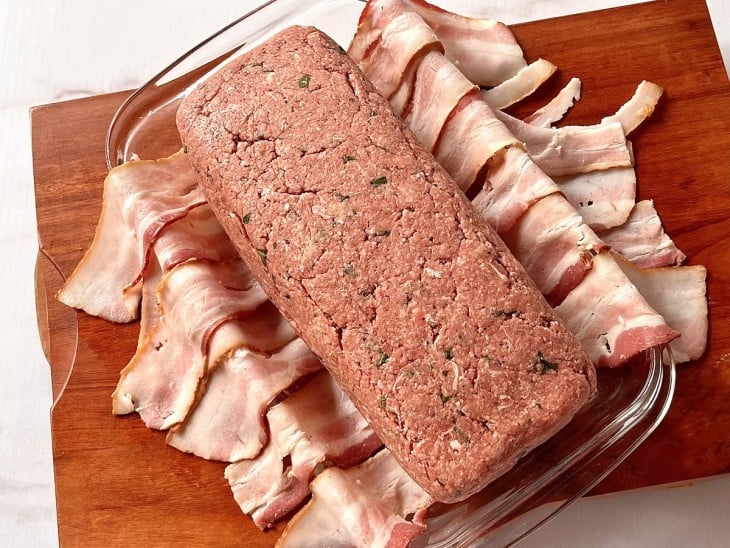 Um refratário forrado com faias de bacon contendo o rocambole de carne.
