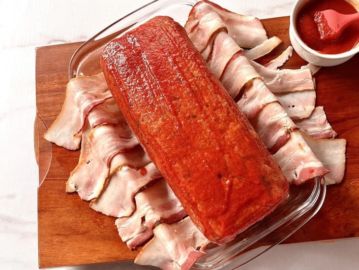 Um refratário forrado com faias de bacon contendo o rocambole de carne pincelado com molho de tomate.