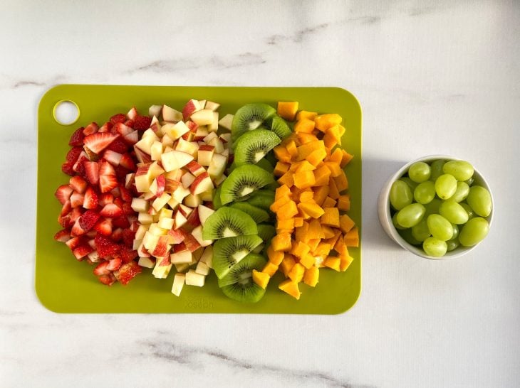 Uma tábua com uva, morango, maçã, kiwi e manga picados.