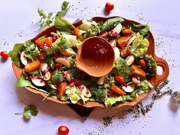 Uma travessa de cor alaranjada forrada com as folhas verdes com um recipiente ao meio para colocar o molho, junto do restante dos ingredientes da salada.