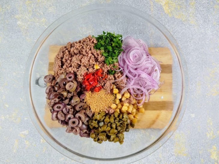 Tigela com grão-de-bico, azeitonas pretas, atum, cebolinha, cebola roxa, mostarda, pimenta, alcaparras, alho e o suco dos limões.
