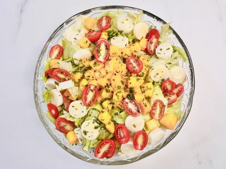Tomates, manga, palmito e cebolinha espalhados sobre a camada de alface dentro da tigela.