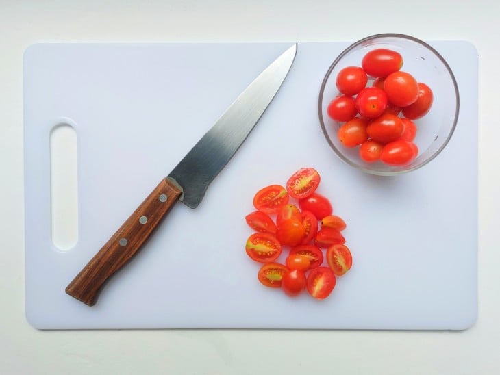 Uma porção de tomate-cereja cortado ao meio e outra porção de tomates-cerejas inteiros dentro de um recipiente. Ambos dispostos em cima de uma tábua na cor branca com uma faca ao lado.