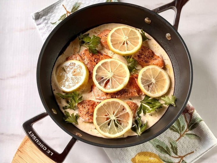 Uma frigideira contendo postas de salmão com molho de creme de leite e rodelas de limão.