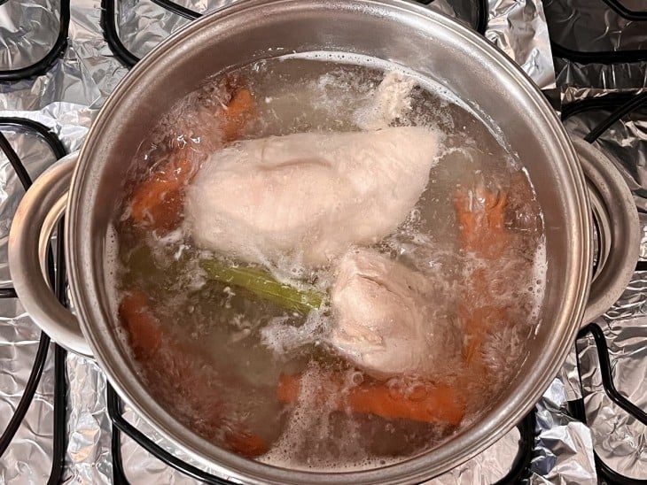 Uma panela contendo peito de frango, talo de salsão, cebola, cenoura e água fervendo.