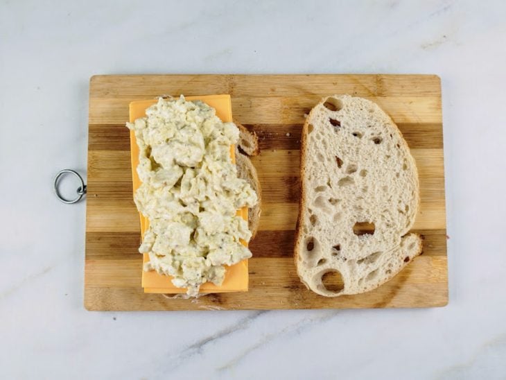 Fatias de pão sobre uma tábua, sendo que uma delas está com fatias de queijo cheddar e ovo sobrepostos.