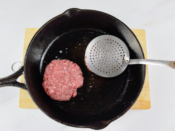Bolinha de carne colocada na frigideira e pressionada por uma escumadeira.