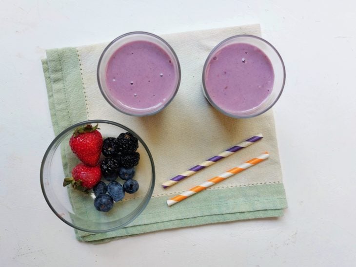 Dois copos com smoothie, dois canudos e algumas frutas congeladas ao lado.