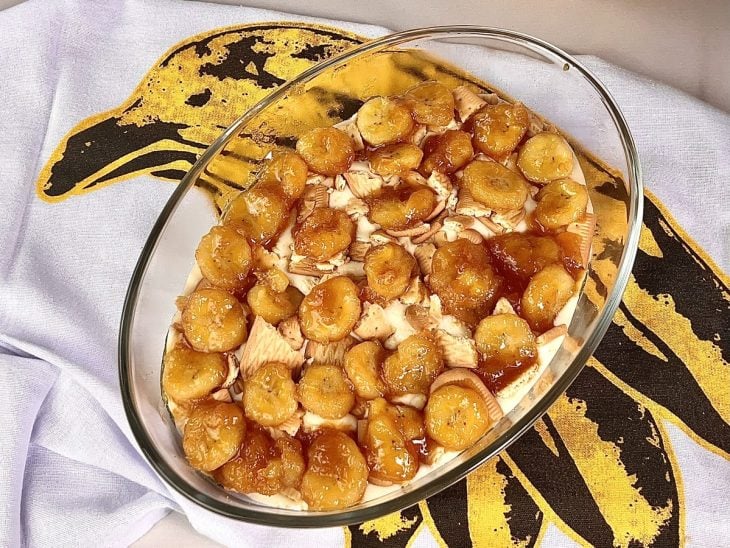 Uma travessa forrada com creme branco doce, coberta com pedaços de bolachas e banana caramelizada.