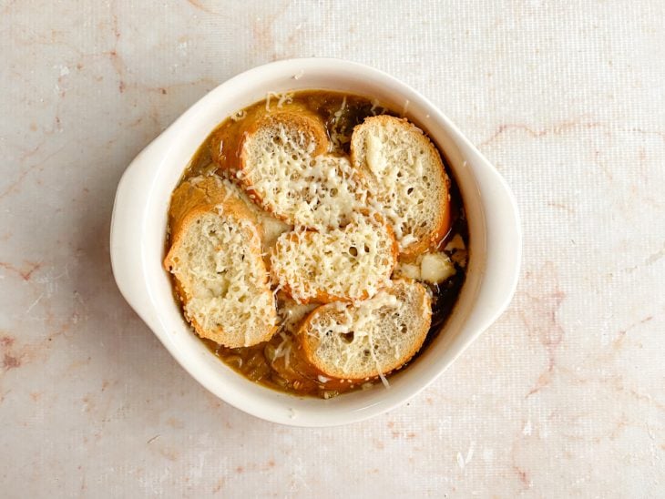 Sopa de cebola, fatias de pão e queijo parmesão ralado em uma tigela.
