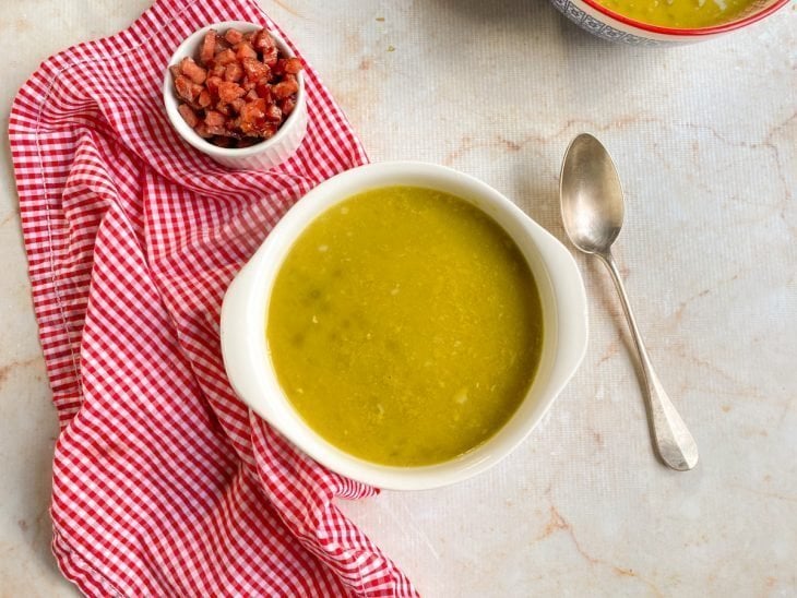 Sopa de ervilha pronta para consumo em um prato fundo ao lado de um potinho com bacon em cubos.