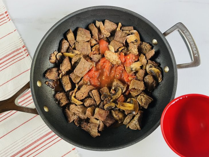 Uma panela com carne, cogumelos, cebola, alho, vinho e extrato de tomate ao fogo.