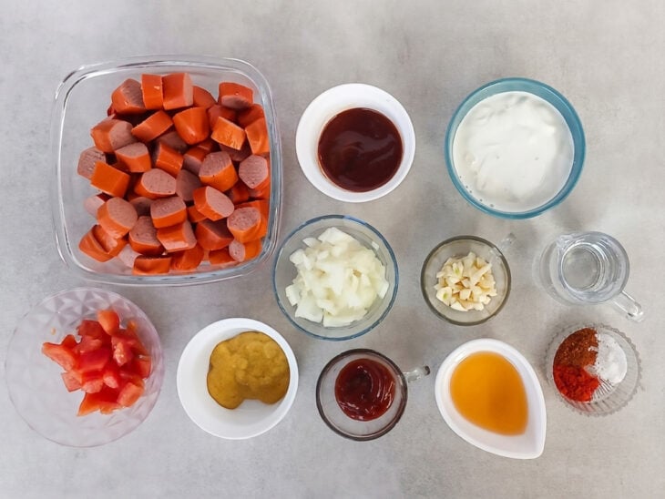 Bancada organizada com salsichas fatiadas em rodelas, tomate, cebola e alho picados em recipientes.
