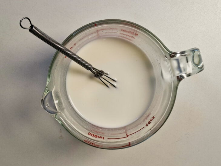 Creme de ricota e água sendo misturados com uma colher dentro de um copo de vidro.