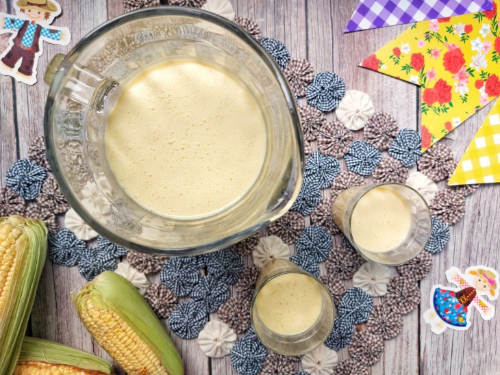 Suco de milho servido em uma jarra e dois copos, dispostos em um mesa com decoração de festa junina.