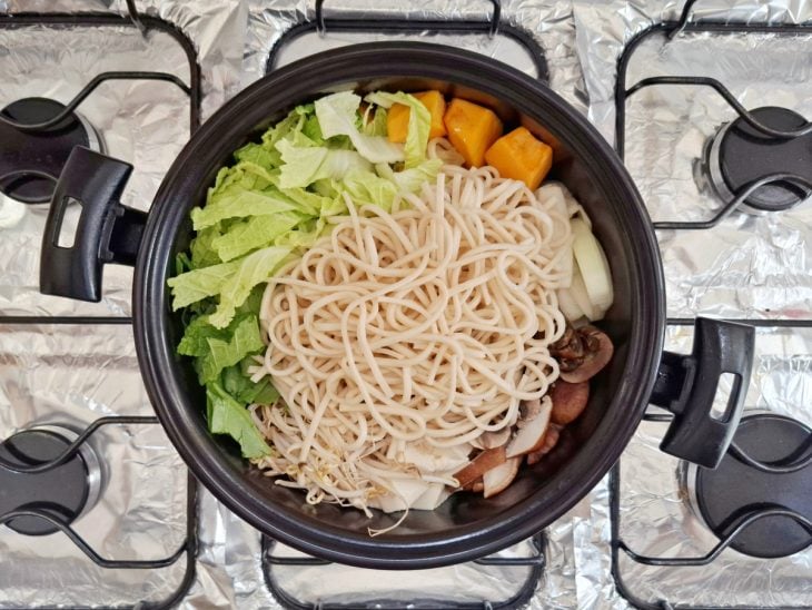 Uma panela com carne, abóbora, cebola, cogumelo, tofu, moyashi, espinafre, acelga e macarrão.