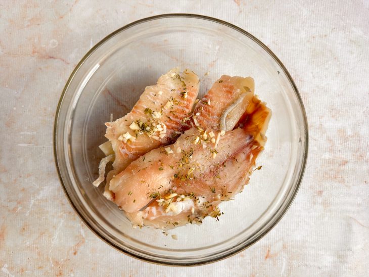 Filés de tilápia sendo temperados com alho, ervas e sal em um recipiente.