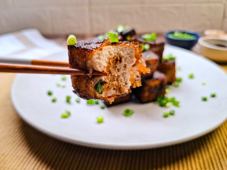 Tofus prontos, servidos em prato, em primeiro plano, um pedaço sendo segurando com um hashi.