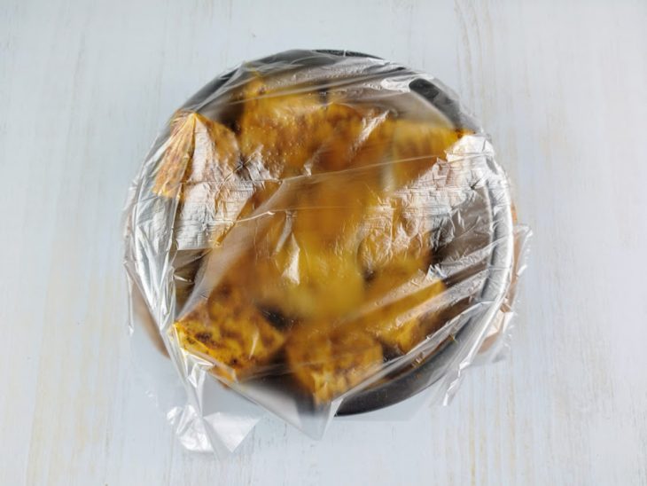 Pedaços de tofu temperados em uma tigela coberta com saquinho plástico de cozinha.