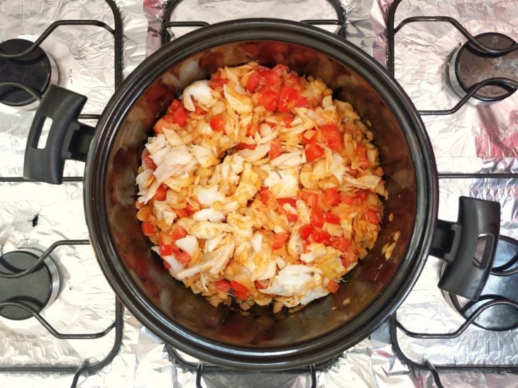 Uma panela refogando alho, cebola, temperos, tomate e bacalhau.