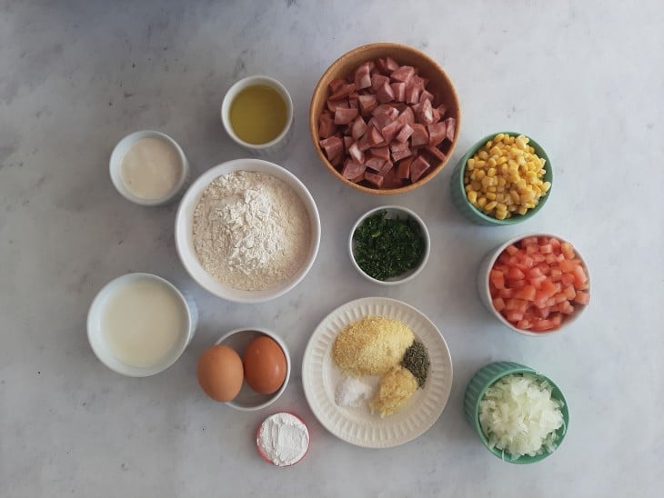 Ingredientes reunidos em diferentes recipientes: calabresa, milho, farinha, cebola. alho, milho, ovos, óleo, leite, farinha, sal, fermento, parmesão, orégano e salsinha.