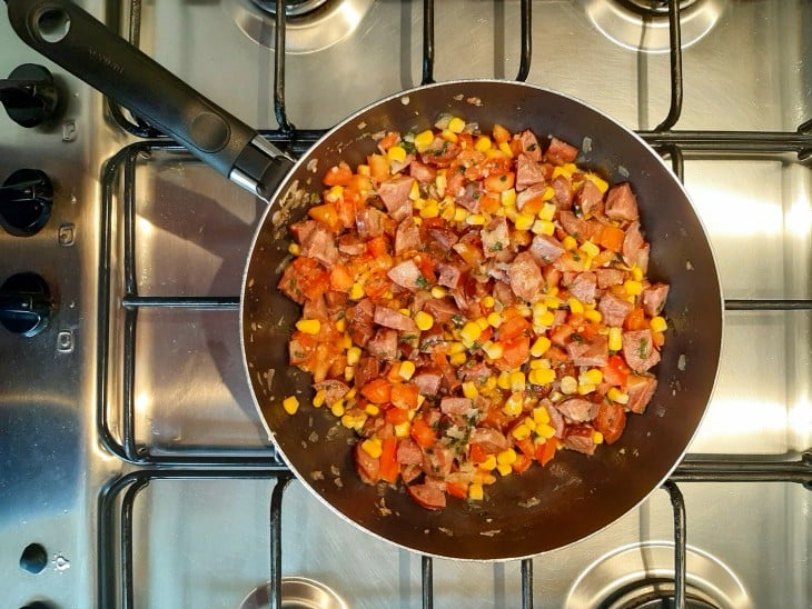 Frigideira com os ingredientes misturados: calabresa, alho, cebola, milho, tomate e salsinha.