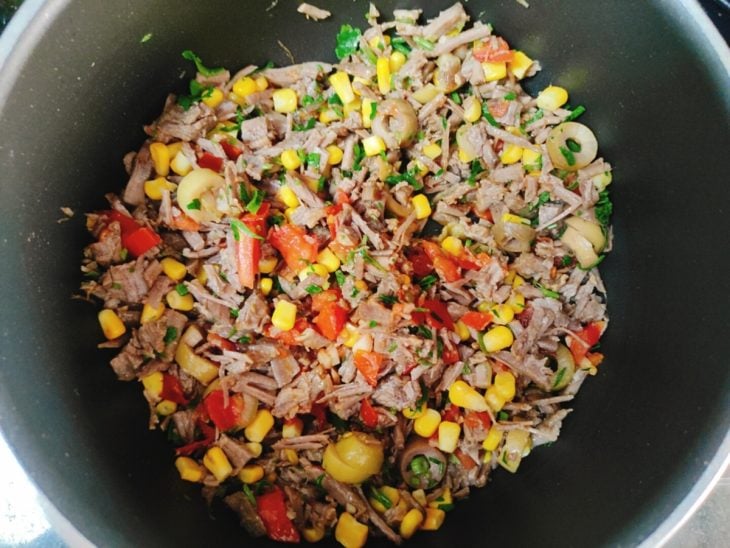 Uma panela com azeite, tomate, alho, carne, pimentão, azeitonas, cheiro-verde, milho.