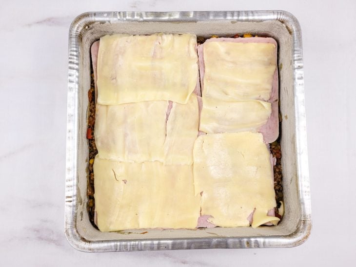 Uma forma forrada com massa da torta e coberta com recheio de carne moída, fatias de queijo e presunto.
