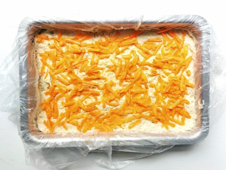 Uma forma retangular com camadas de pão, frango desfiado cremoso e cenoura ralada.