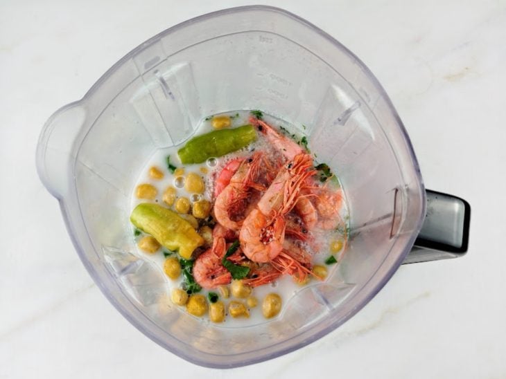 Um liquidificador com tomates, cebola, alho, coentro, cebolinha, amendoim, pimentas-de-cheiro, gengibre, leite de coco, sal, água e camarão.